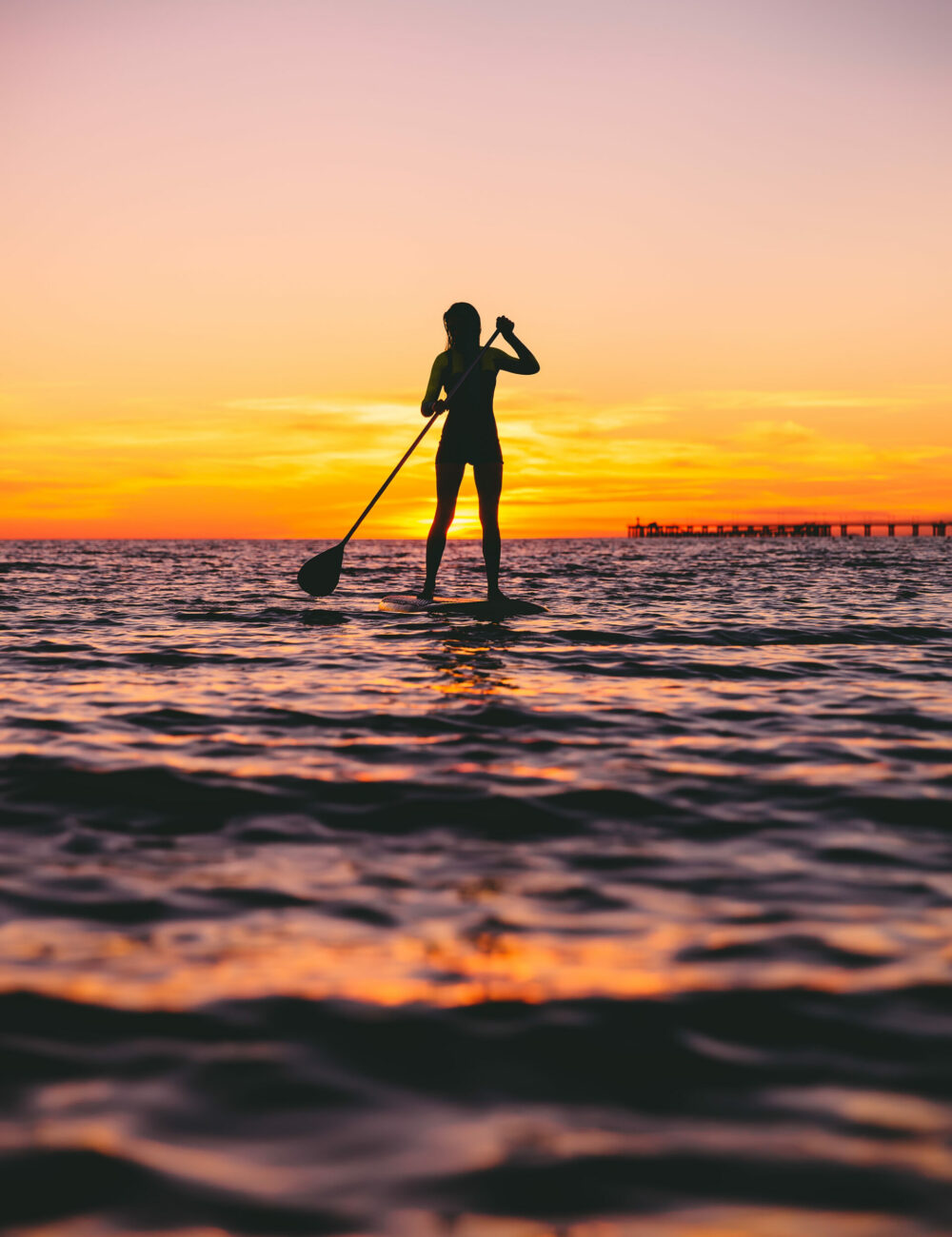 Woman stand up paddle boarding at dusk on a flat warm quiet sea with beautiful sunset colors